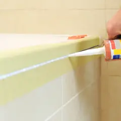 Ręka z pistoletem wyciska biały silikon, uszczelniając połączenie wanny z płytkami.