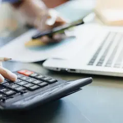 Dłoń z ołówkiem liczy na kalkulatorze, obok laptop i kubek. Analiza rachunków, może zużycie kwh.