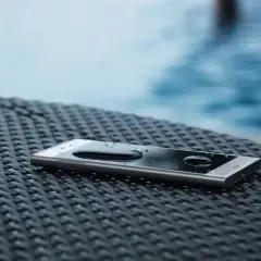 Telefon z certyfikatem **IP65** leży na stoliku przy basenie, krople wody na ekranie świadczą o jego wodoodporności.