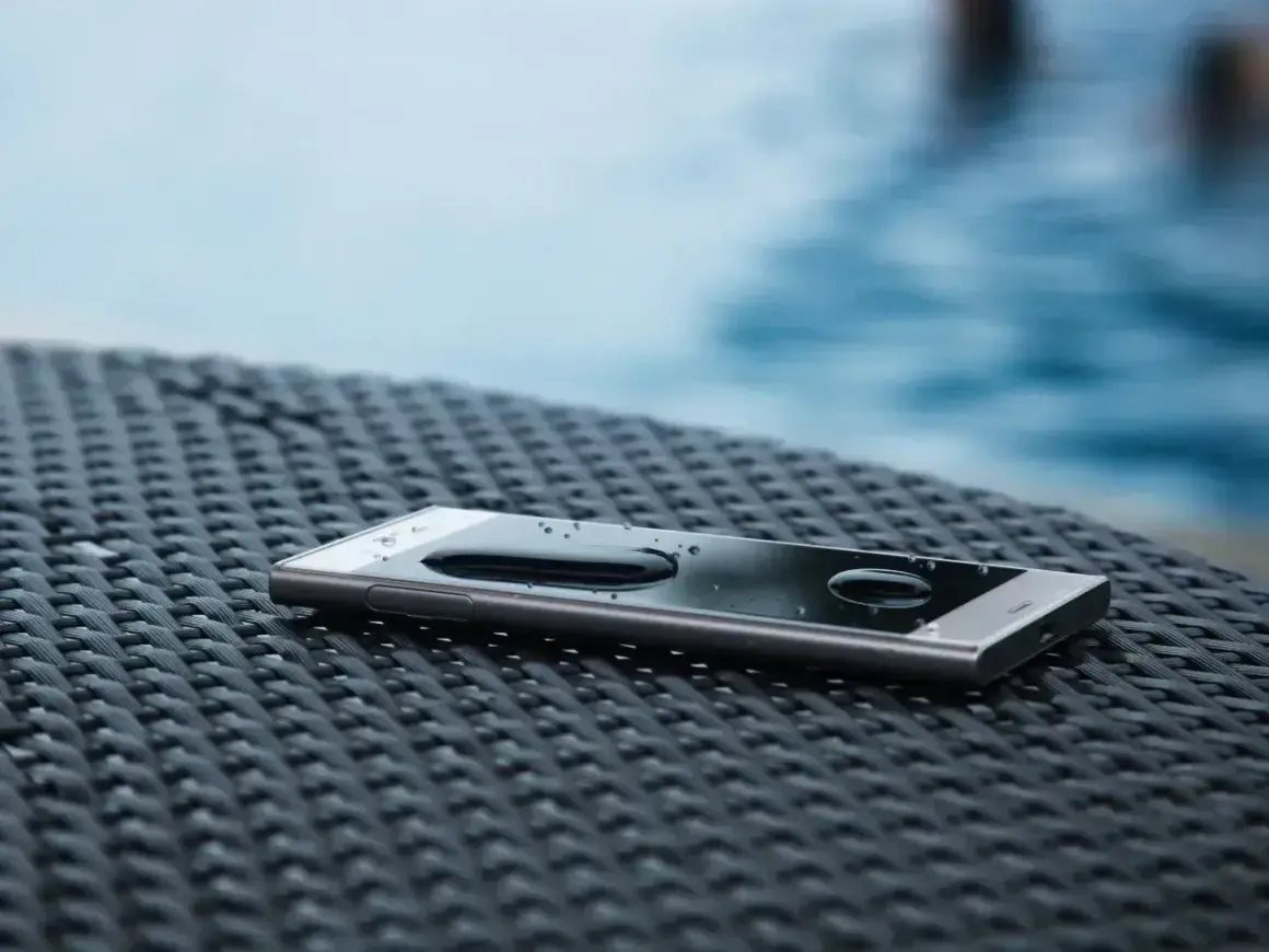 Telefon z certyfikatem **IP65** leży na stoliku przy basenie, krople wody na ekranie świadczą o jego wodoodporności.