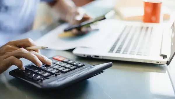 Dłoń z ołówkiem liczy na kalkulatorze, obok laptop i kubek. Analiza rachunków, może zużycie kwh.