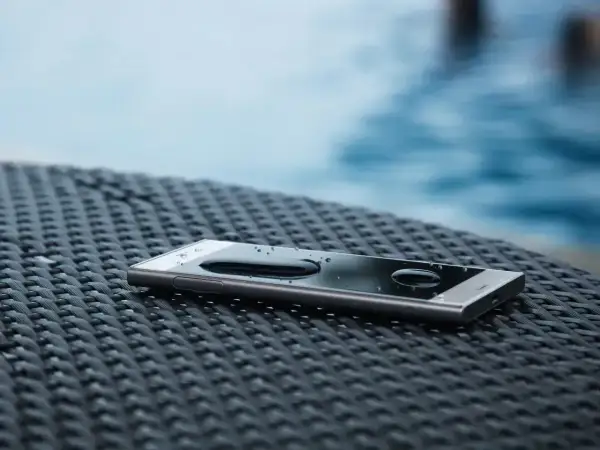 Telefon z certyfikatem **IP65** leży na stoliku przy basenie, krople wody na ekranie świadczą o jego wodoodporności.